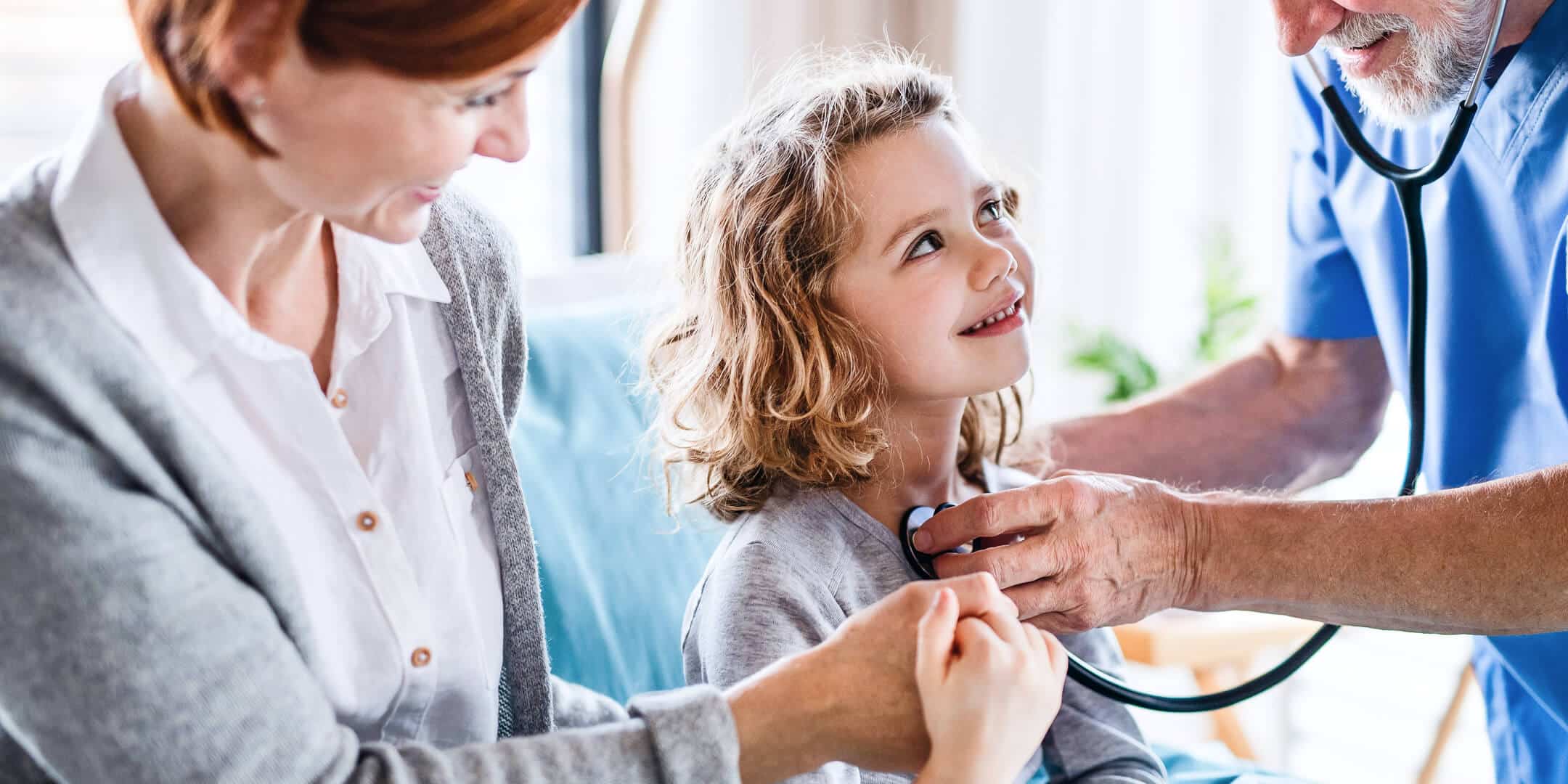 Nuori tyttö istuu äitinsä sylissä lääkärin vastaanotolla ja hymyilee