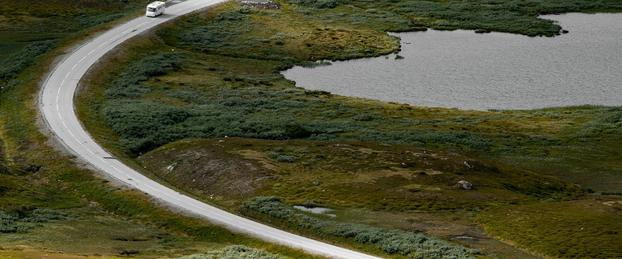 Asfalttitie mutkittelee vihreässä maisemassa lähellä järveä, tiellä kulkee valkoinen asuntoauto.