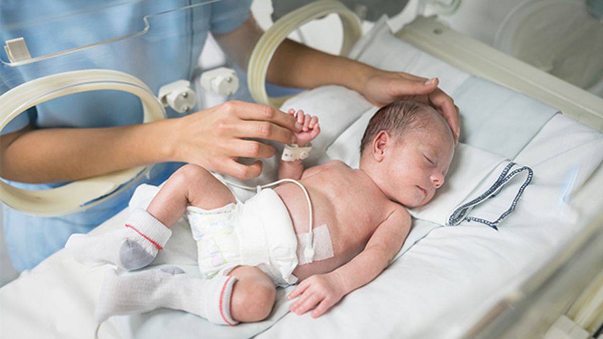 Keskosvauva makaa inkubaattorissa, hoitaja koskettaa vauvaa hellästi kädestä.