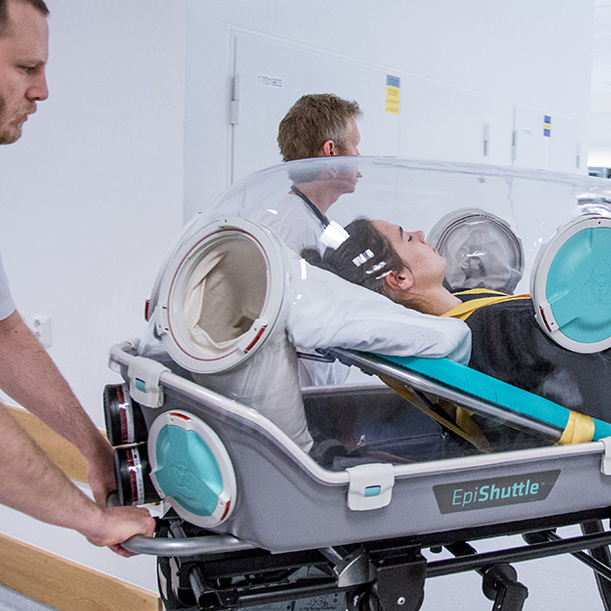 En patient ligger i en genomskinlig isoleringsbår märkt EpiShuttle, eskorterad av två vårdpersonal i en sjukhuskorridor.