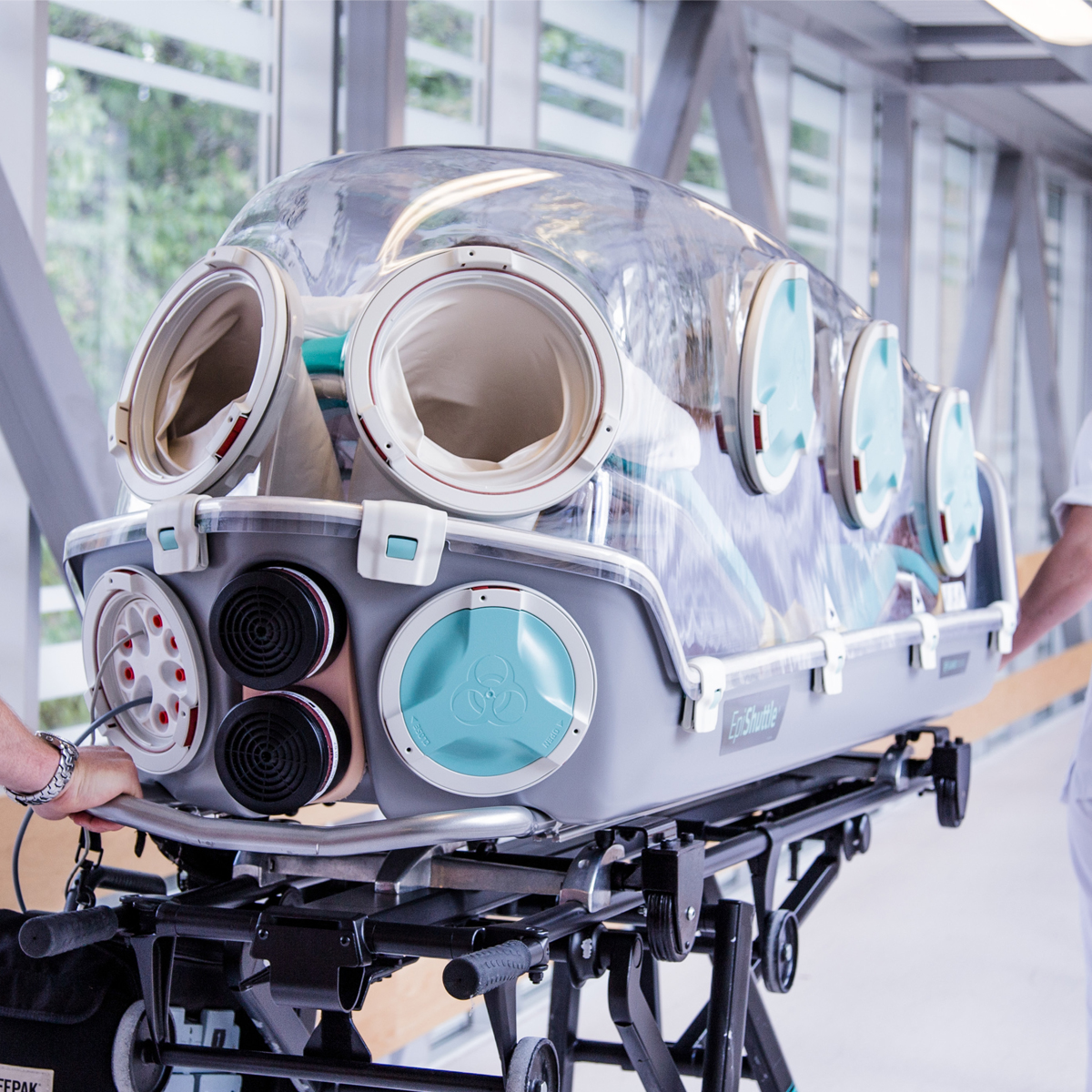 Medicinsk isoleringstransportsäck med transparent lock och armportar i en sjukhuskorridor.