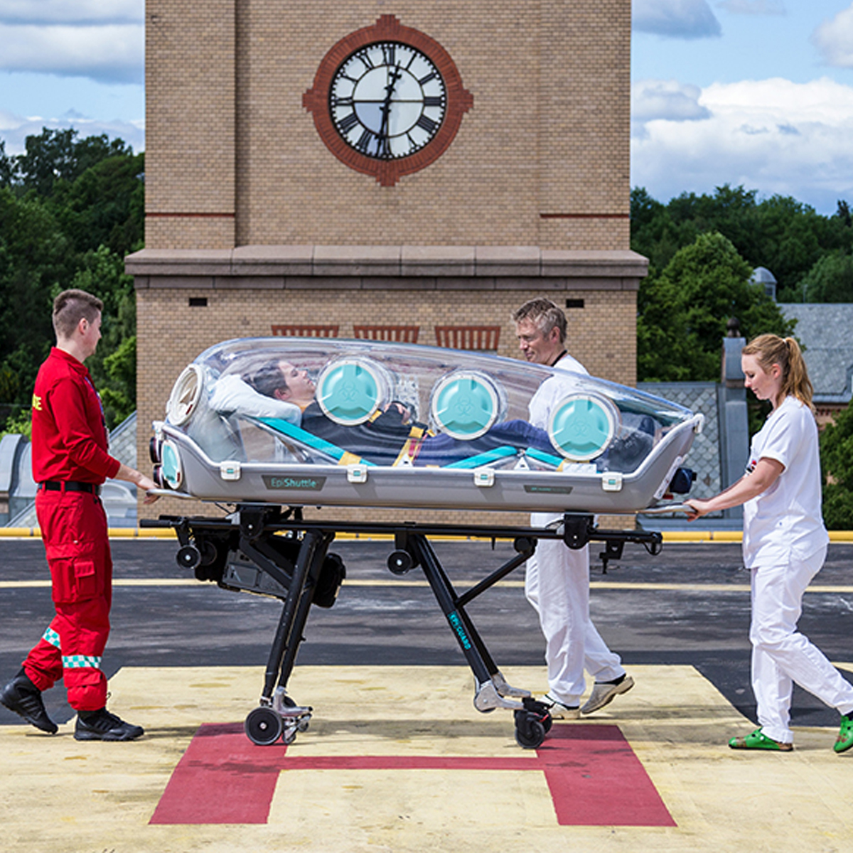 Tre medicinska personal transporterar en patient i en hjulburen isoleringskapsel på ett sjukhustak för helikoptrar.