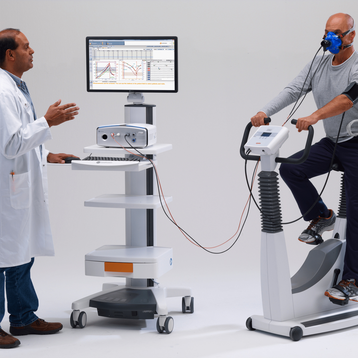 Henkilö polkee kuntopyörää maski kasvoilla, toinen henkilö laboratoriotakissa ohjaa testiä tietokoneella.