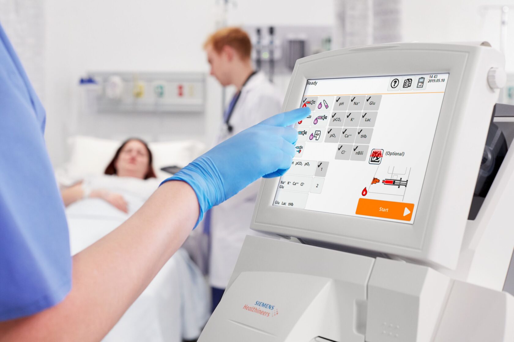 Hoitaja käyttää lääketieteellistä laitetta kosketusnäytöllä, taustalla potilas sairaalasängyssä.