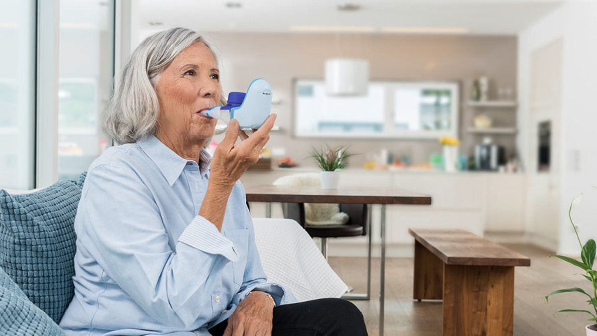 Vanhempi nainen istuu sohvalla käyttäen sinistä hengitysilmapuhallinta kirkkaassa modernissa keittiössä.