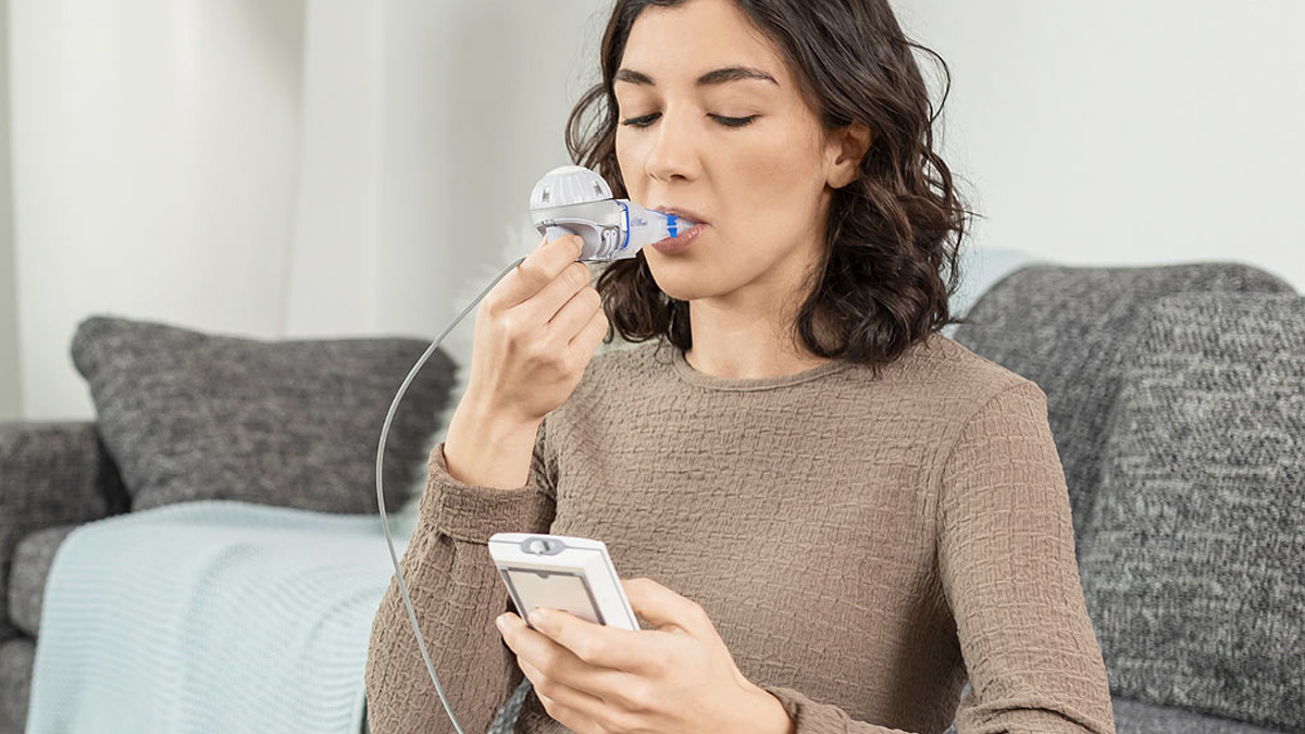 Nainen käyttää kannettavaa hengityslaiteetta istuessaan sohvalla ja pitää kädessään yhdistettyä mobiililaitetta.