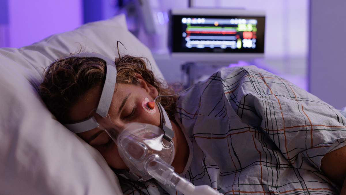 Patient i sjukhussäng med syrgasmask, medicinsk monitor i bakgrunden som visar vitala tecken.