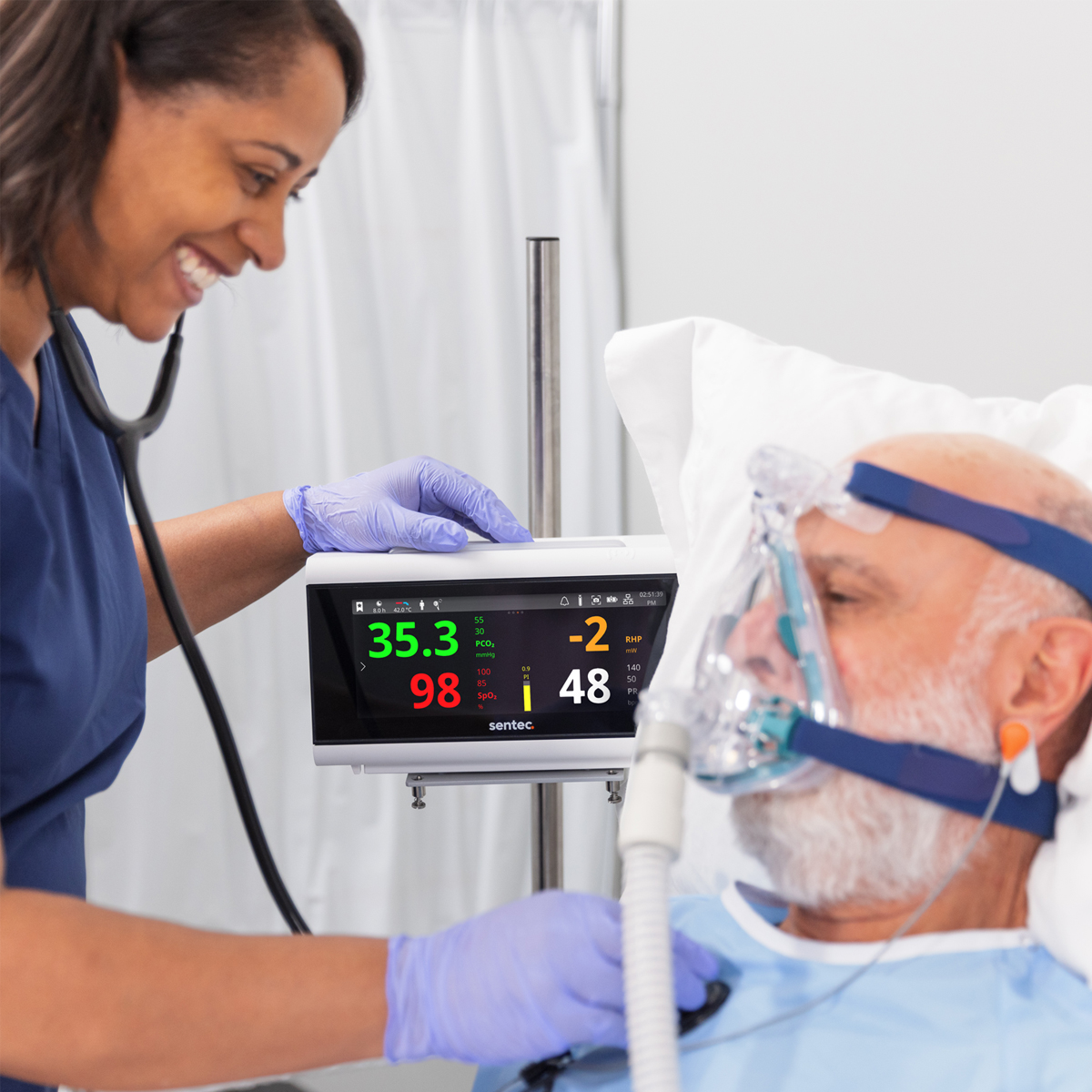 Sjuksköterska kontrollerar vitala tecken på monitor för äldre manlig patient som bär syrgasmask i sjukhussäng.