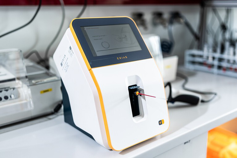 Valkoinen laboratoriolaite, jossa on kosketusnäyttö, pöydällä laboratoriossa.