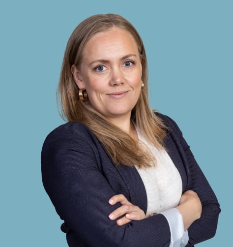 Pitkähiuksinen nainen, jolla on laivastonsininen bleiseri ja valkoinen paita, kädet ristissä, sinistä taustaa vasten.