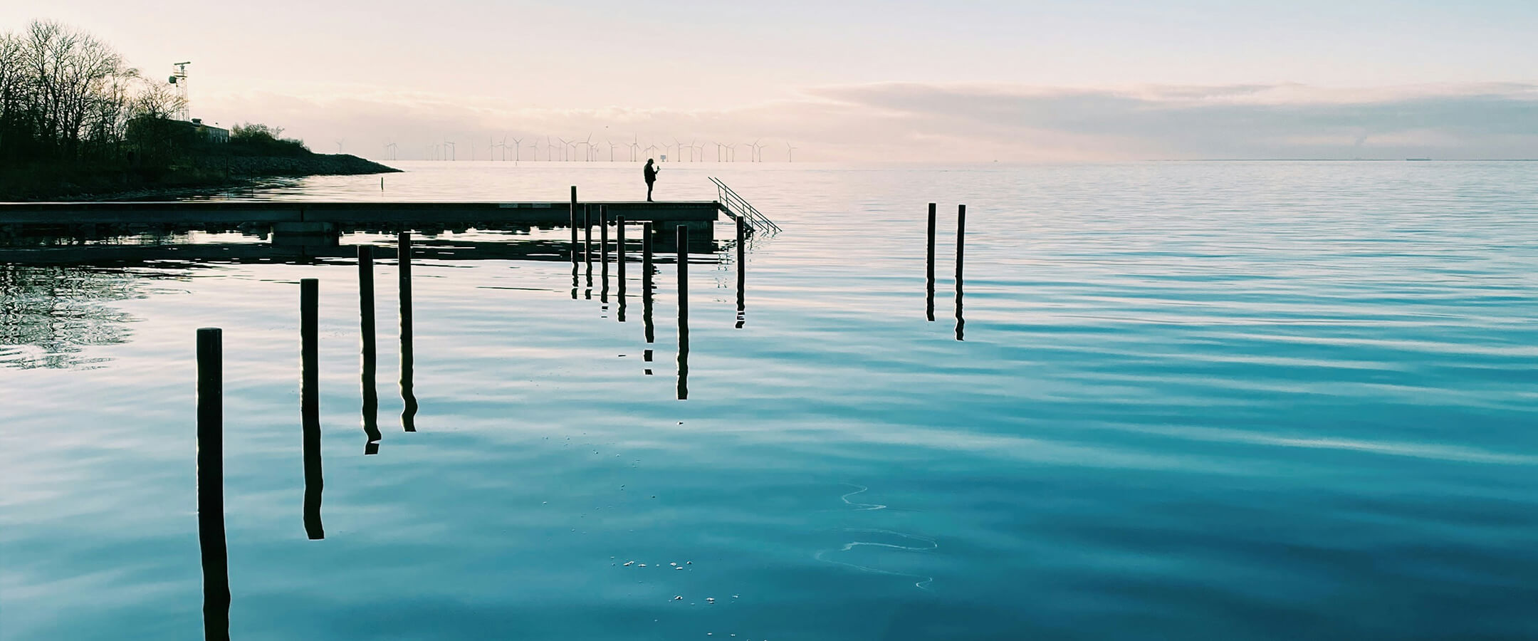Henkilö seisoo laiturilla tyynen veden yllä auringonlaskussa, kaukana horisontissa näkyy tuulivoimaloita.