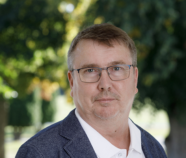 Muotokuva silmälasipäisestä miehestä tummassa takissa vehreää taustaa vasten.