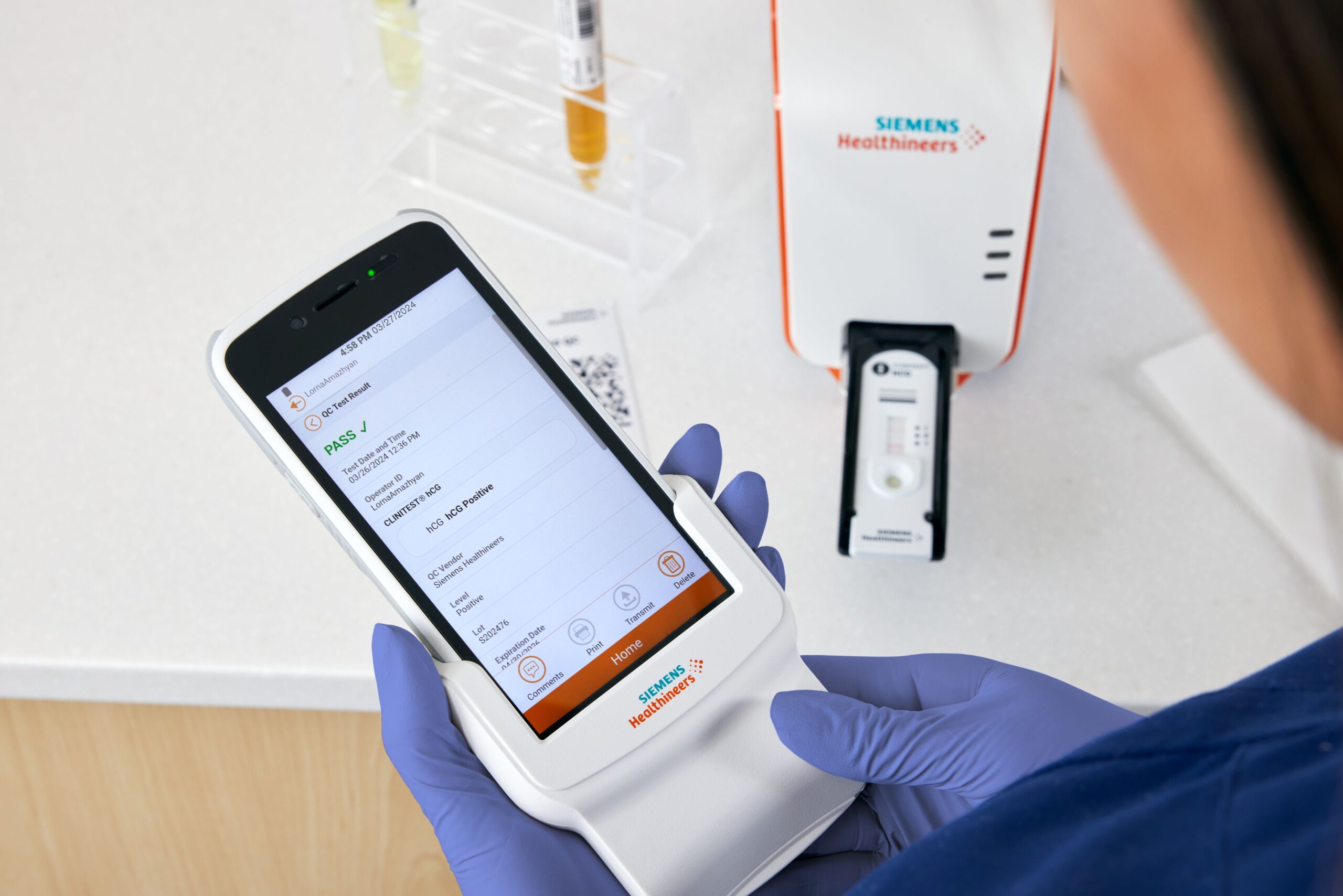 Hansikkaisiin pukeutuneet kädet pitävät Siemens Healthineersin diagnostista laitetta, joka näyttää testituloksen lääketieteellisessä laboratorion ympäristössä.